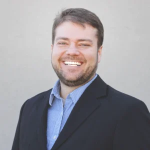 This image shows a professional headshot of a smiling man in formal attire, wearing a dark blazer over a light blue shirt. His approachable and friendly expression makes the portrait ideal for use in business-related settings such as LinkedIn profiles, corporate websites, and professional presentations. The neutral background ensures focus on the subject, highlighting his confident and positive demeanor, perfect for personal branding and networking.