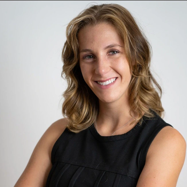 This image features a professional headshot of a smiling woman with light brown wavy hair, dressed in a sleeveless black top. Her approachable demeanor and confident posture make this portrait perfect for use in business settings such as LinkedIn profiles, corporate websites, and networking platforms. The clean, minimal background ensures the focus remains on her professional yet friendly appearance, suitable for showcasing personal branding or professional services.